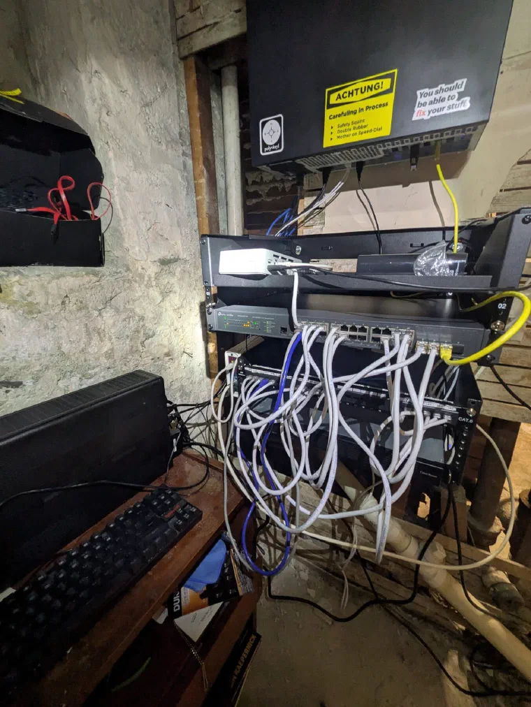 A homelab server rack in a stone-walled basement — a patch panel and network switches with a tangle of blue, white, and yellow ethernet cables, beside a keyboard.