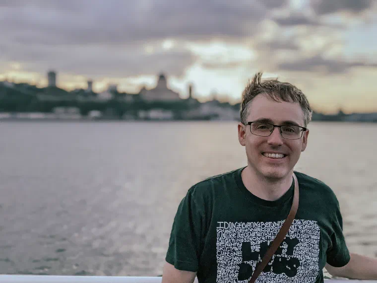 Jared Knowles smiling at the camera at golden hour, a river and a city skyline behind him, a camera strap over one shoulder.